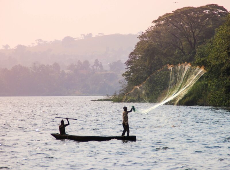 Two fishermen in a boat on a lake, one throwing a wide net, during luxury Uganda holidays.