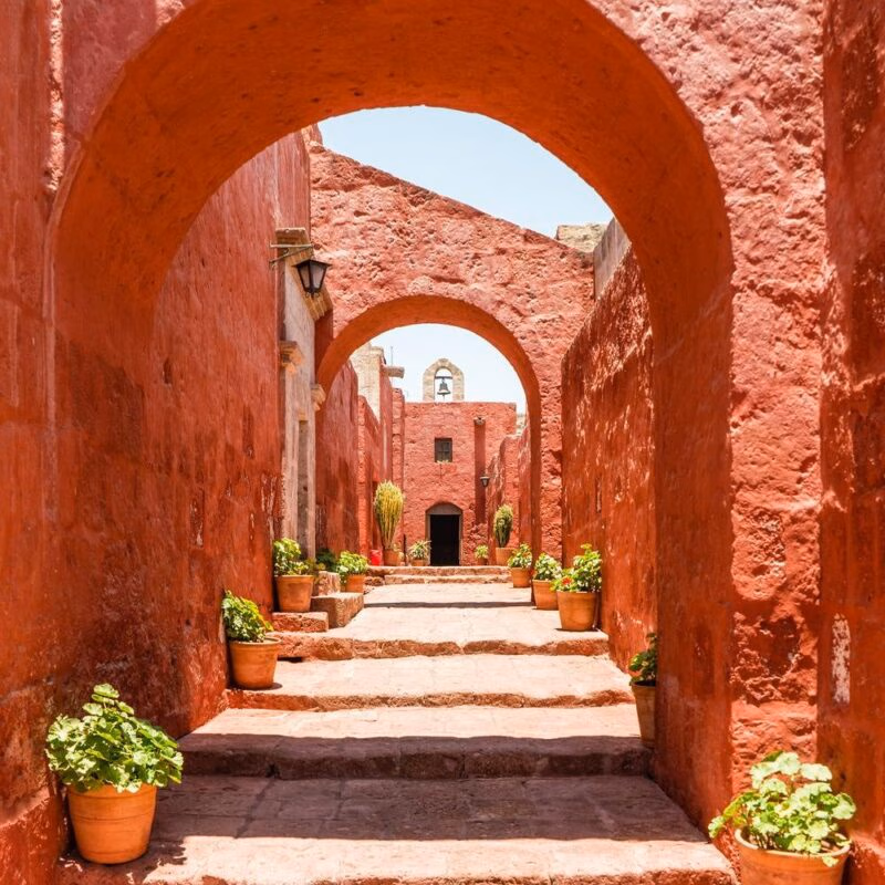 Luxury Peru Tours - Arched pathway in a red-walled monastery with potted plants under bright sunlight.