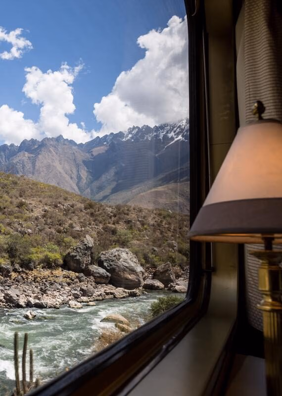 A view from a train window looks out at a rushing river and steep, rocky, scrub-covered mountainside with snow-capped peaks, a great luxury Machu Picchu holidays.