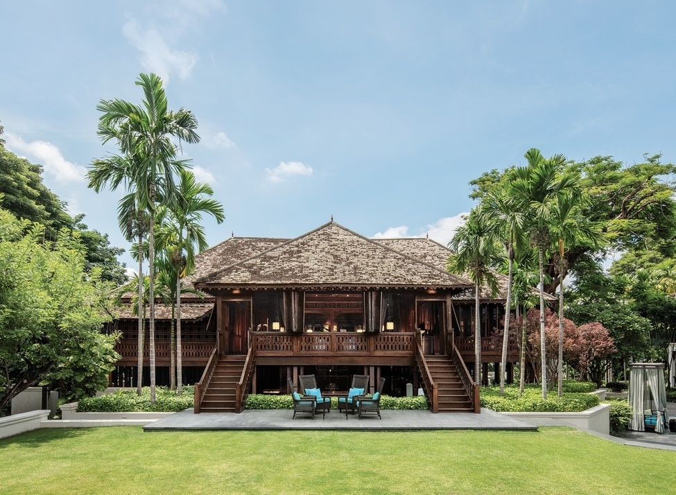 A large, traditional dark wood Thai building with twin staircases and a symmetrical facade surrounded by palm trees.