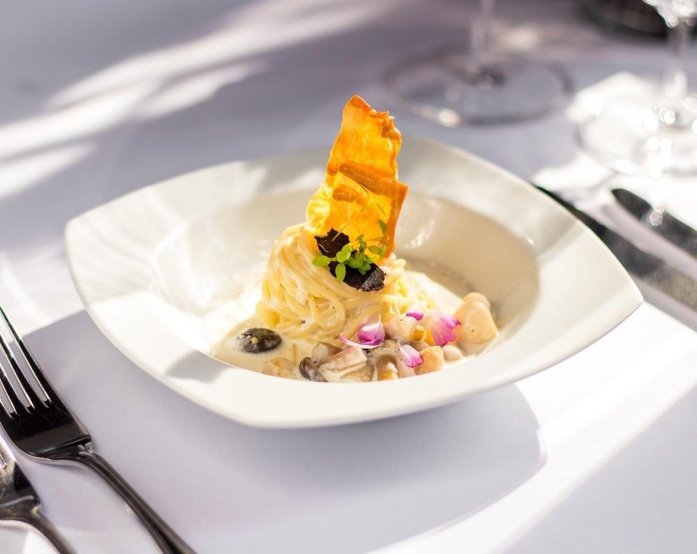 A gourmet pasta dish with cream sauce, a tall golden garnish and pink flowers on a white tablecloth.