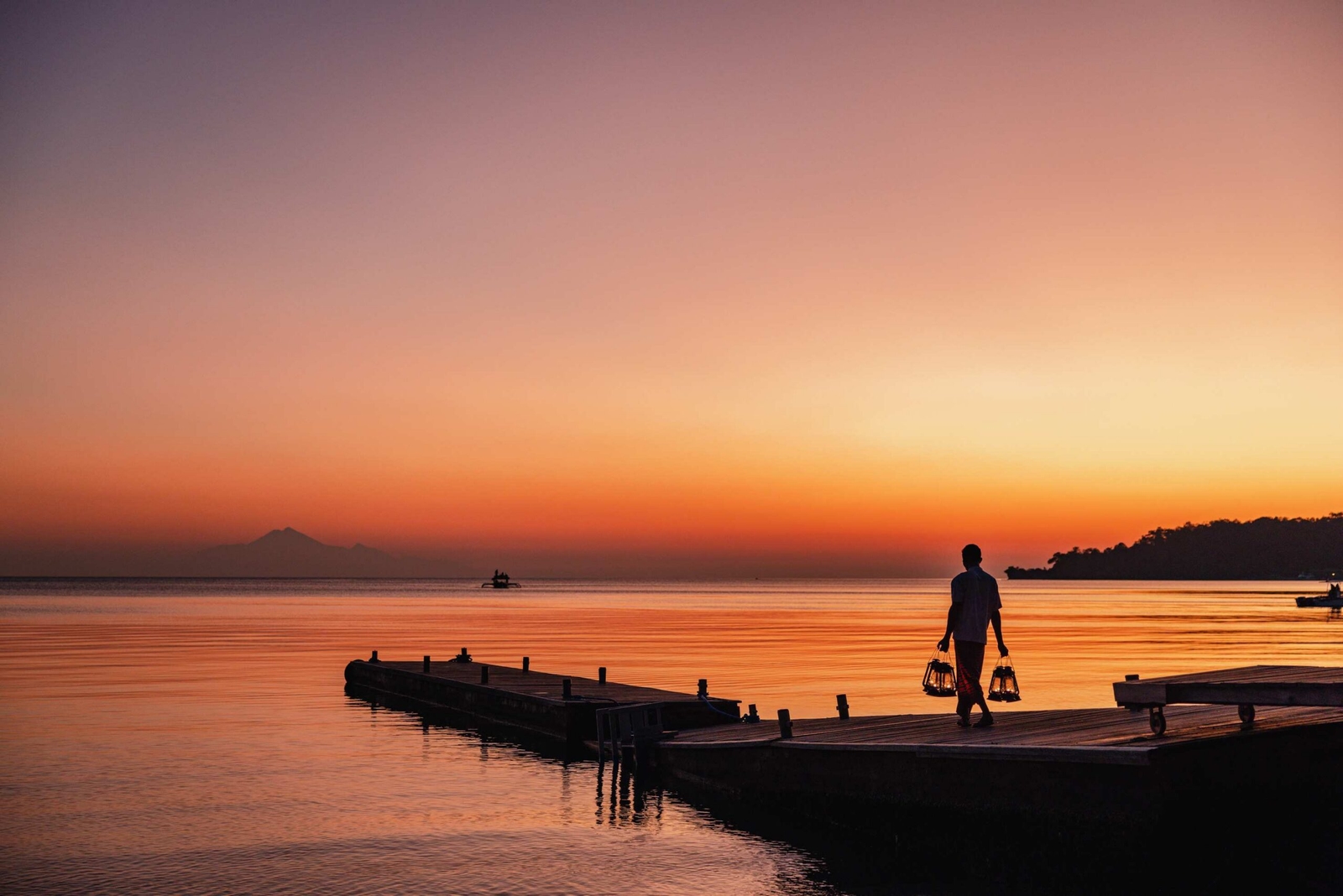 Silhouette of a person walking on a pier carrying glowing lanterns at sunset with mountains in the distance.