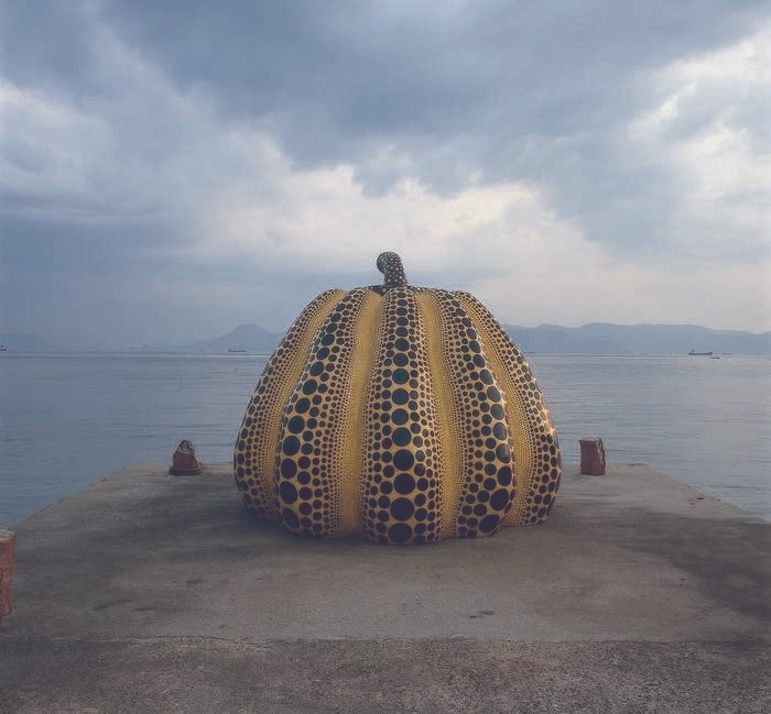 A large yellow and black polka-dotted pumpkin sculpture on a pier with the ocean and mountains in the background.