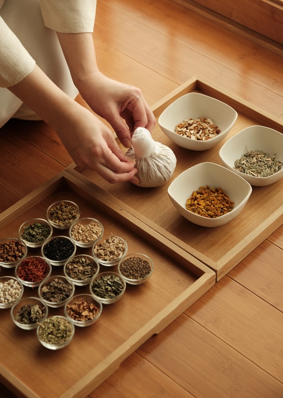 Hands tying a cloth herbal compress at Beniya Mukayu beside a wooden tray containing various dried medicinal herbs.