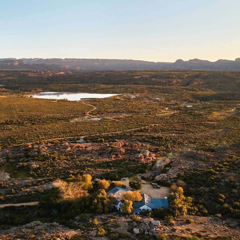 Aerial view of a remote house amid arid terrain at sunset with a lake in the background.