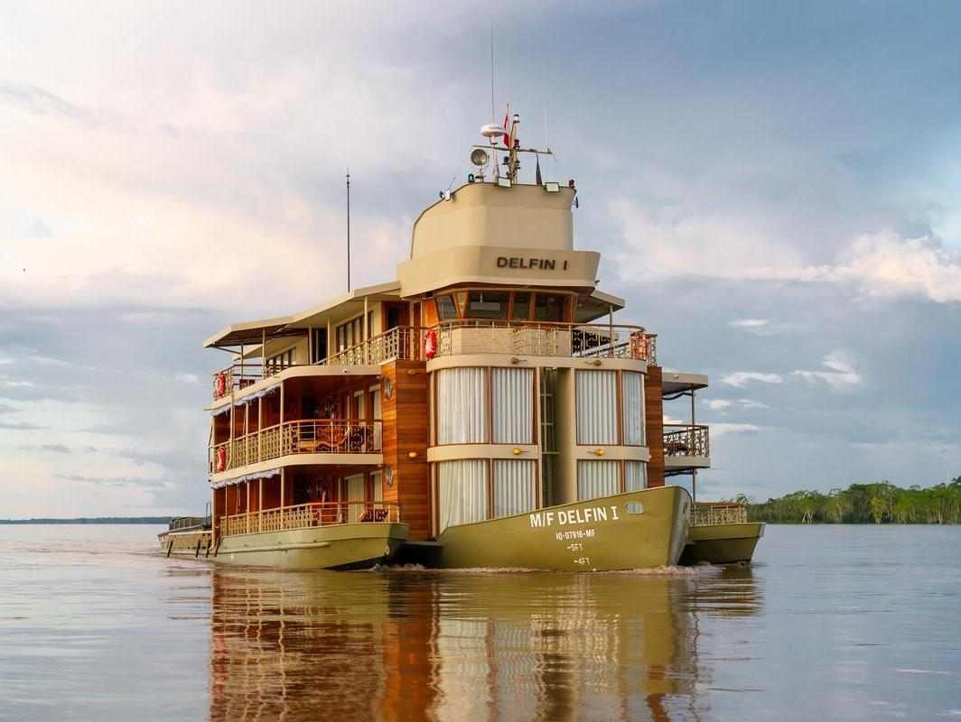 The exterior of Delfin I, a luxury wooden river cruise, sailing on a wide river under a cloudy sky.