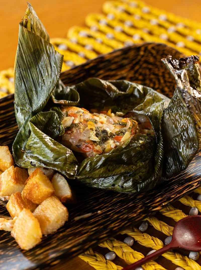 A plated meal of fish steamed in a green leaf with a side of golden roasted potatoes on a dark plate.