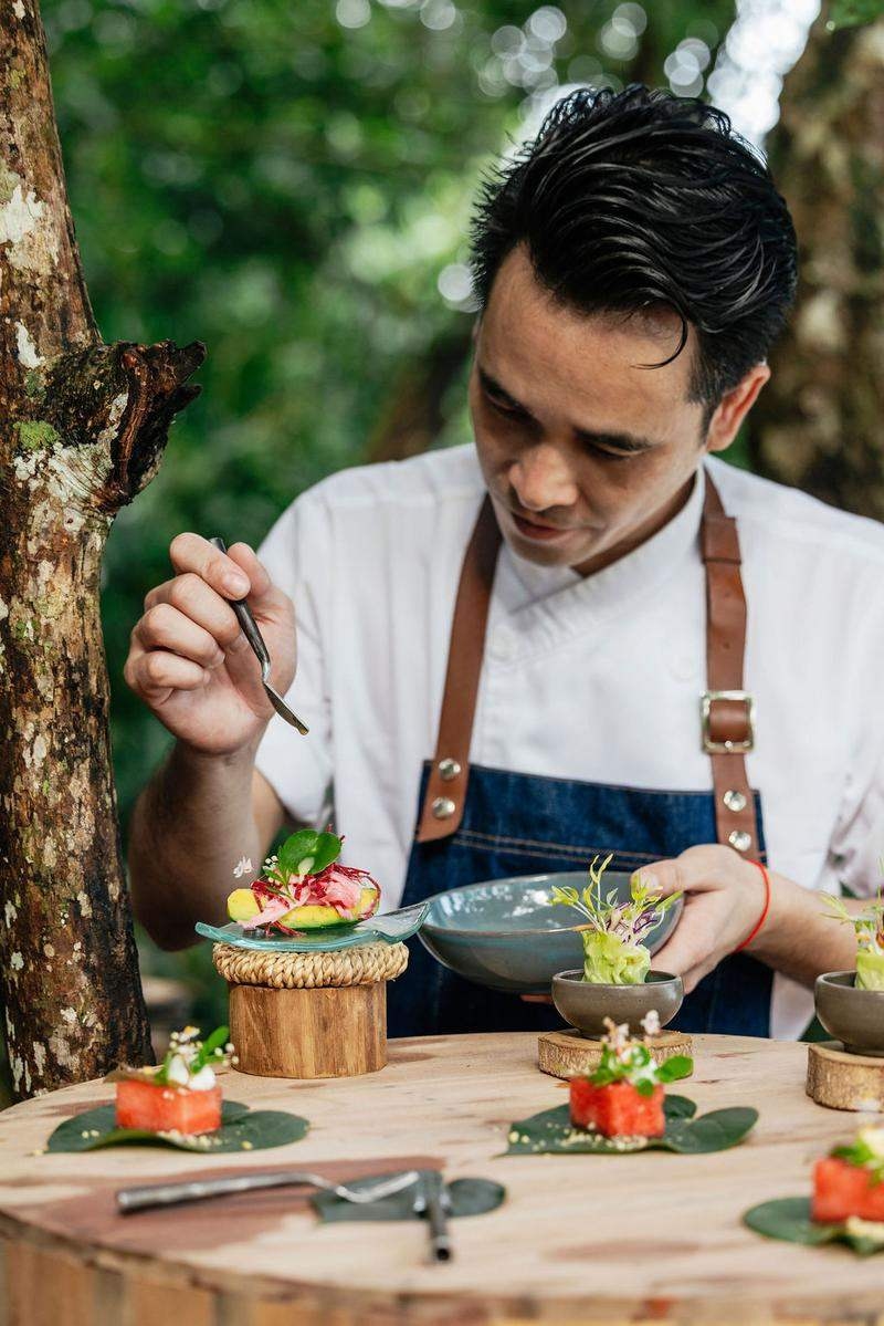 A chef using tweezers to garnish small colourful dishes on a wooden outdoor table.