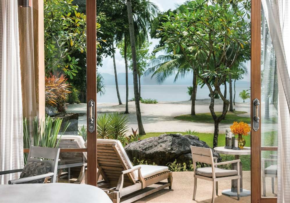 View from a bedroom interior at Four Seasons Langkawi looking out past a dining table and pool toward a white sand beach and ocean.
