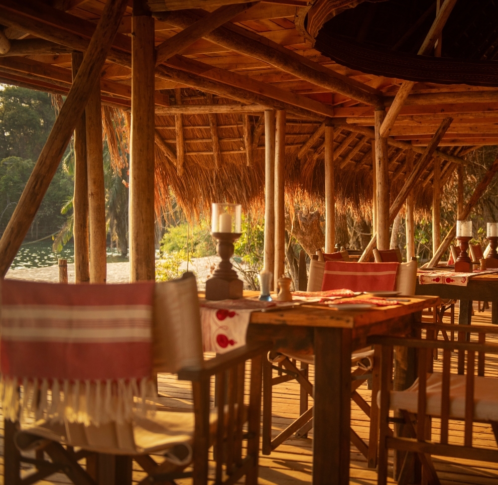 Rustic open-air dining area with thatched roof overlooking a natural landscape at sunset.
