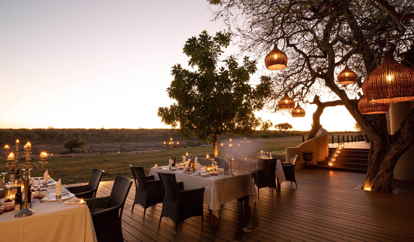 Outdoor dining setup with lit candles on tables, hanging lanterns on a tree at dusk.