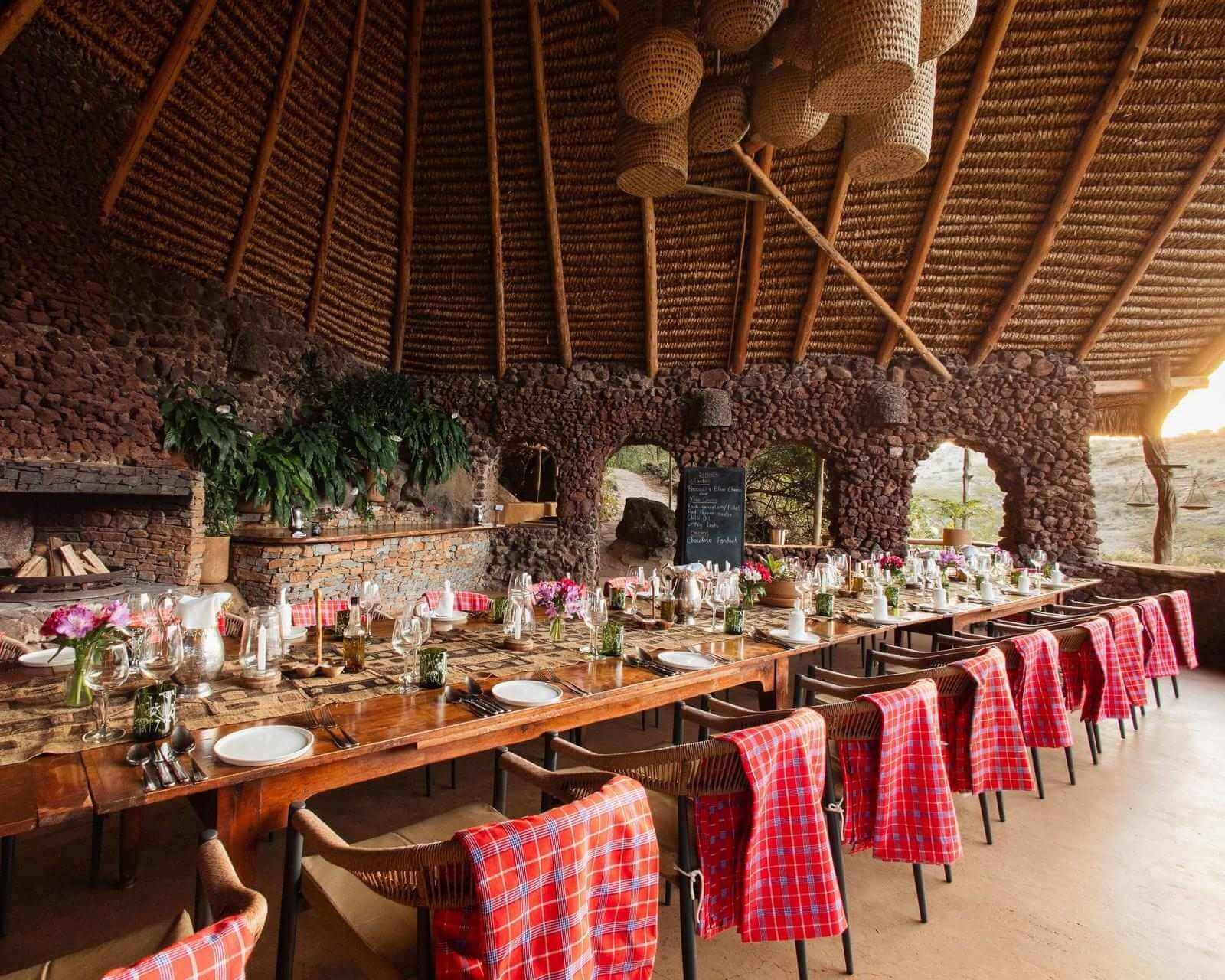 A long set wooden dining table under a large thatched roof in a rustic open-air dining space.