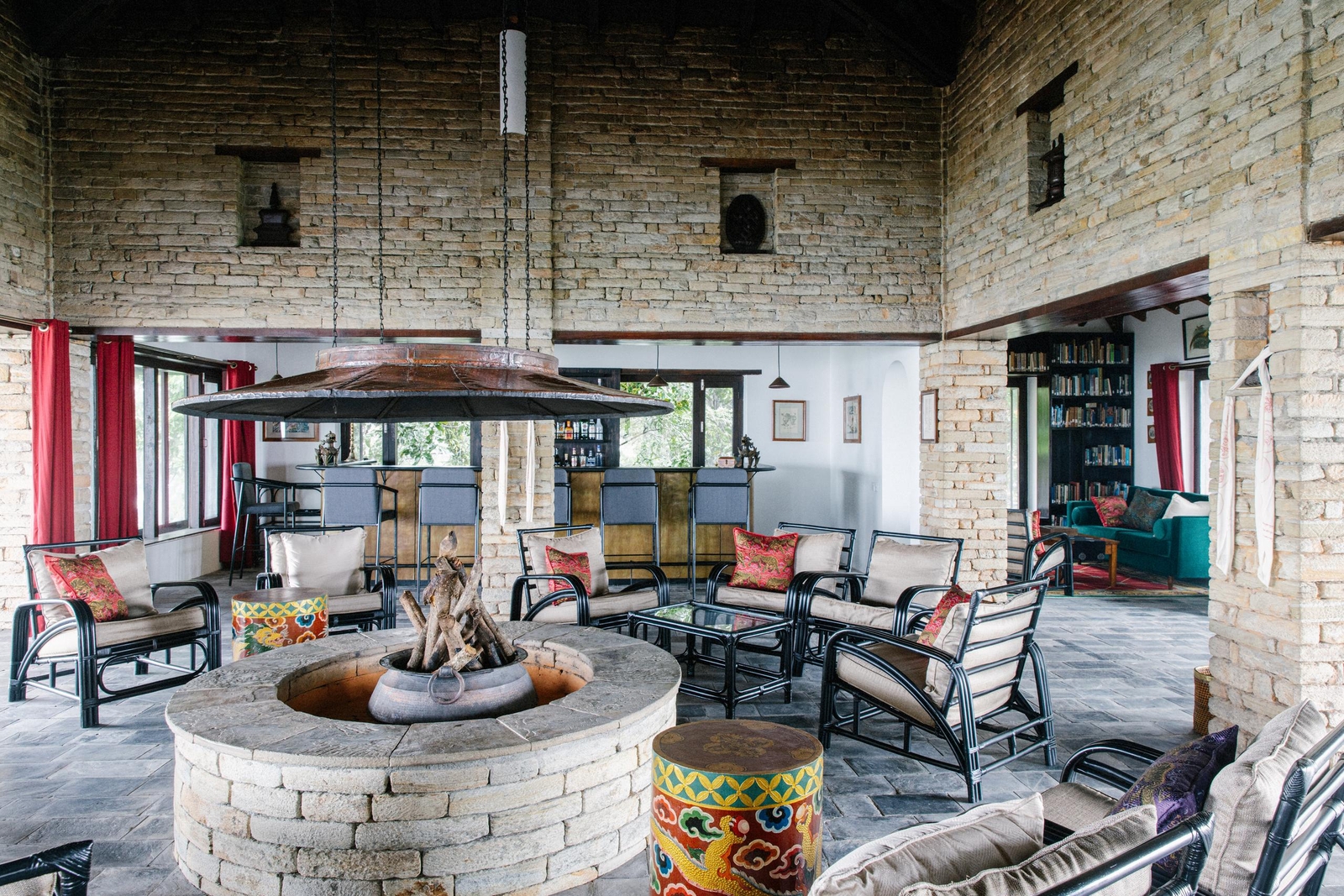 Rustic lounge with brick walls, central fire pit, metal chairs, and colorful accents.