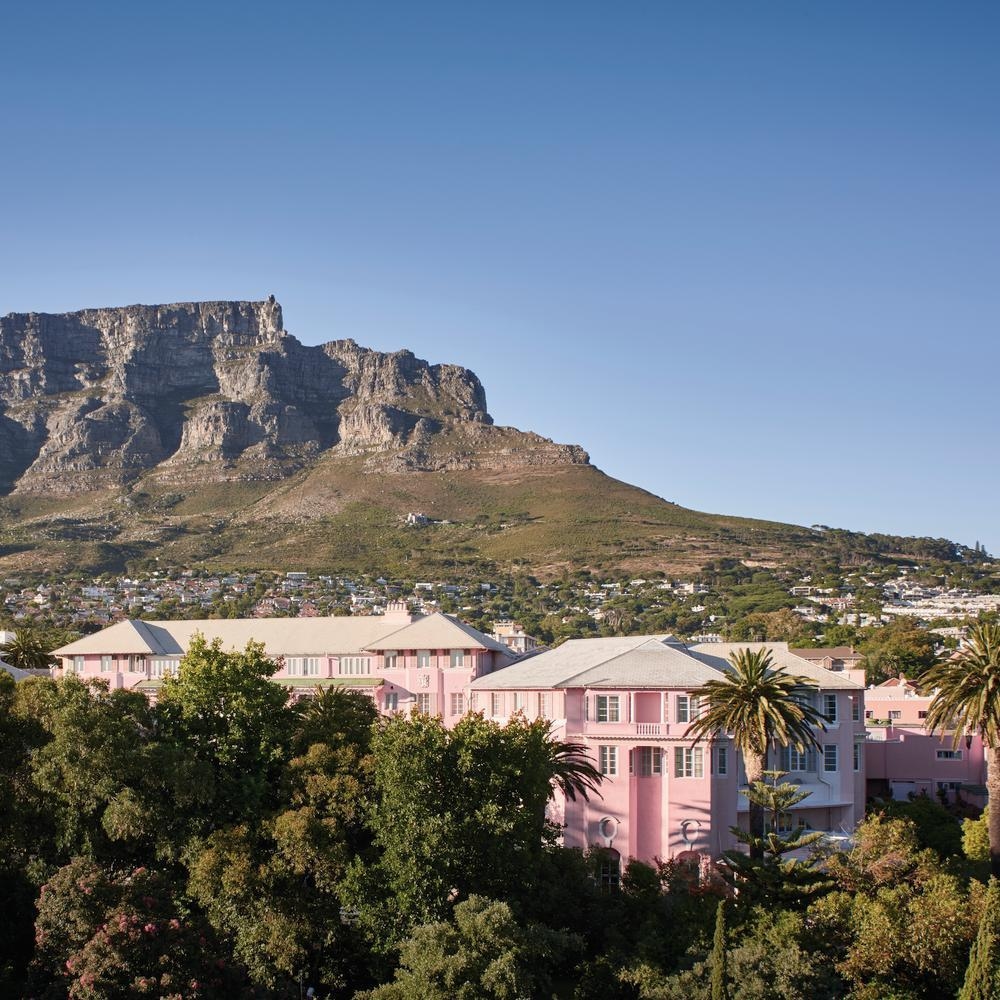 Exterior view of a grand pink hotel surrounded by trees with the flat-topped Table Mountain in the distance.