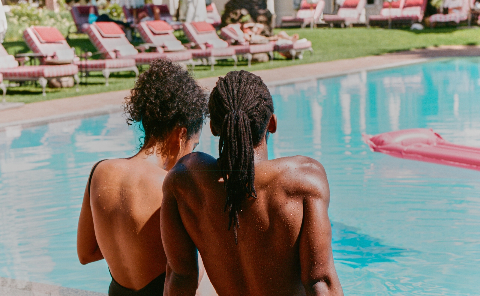 Close-up from behind of two people sitting at the edge of a turquoise swimming pool on a sunny day.