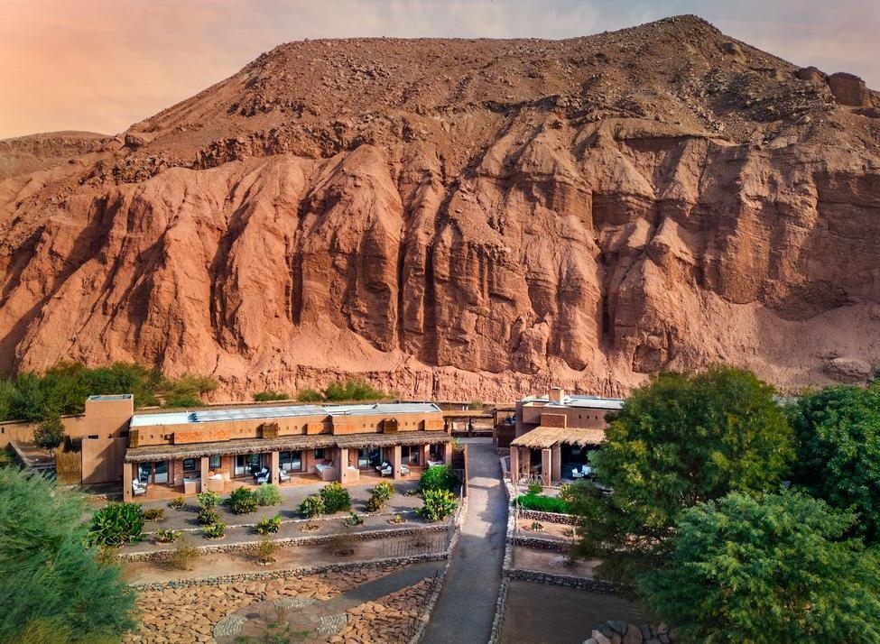 Aerial view of Nayara Alto Atacama with low-rise buildings and greenery nestled directly at the base of large red rock cliffs.