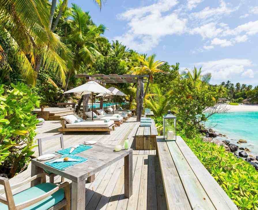 Alfresco dining table and sunbeds on a wooden platform overlooking a tropical beach and clear water.