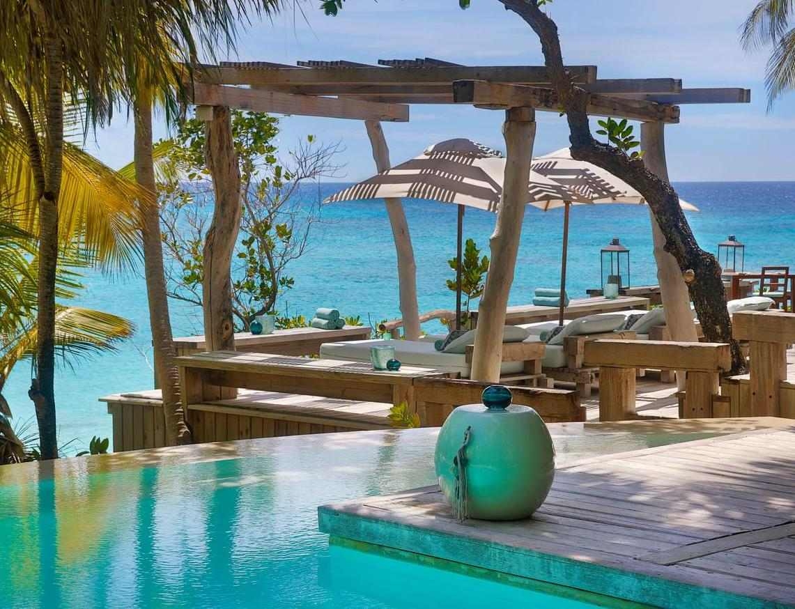 A luxury infinity pool at a tropical resort facing the bright blue sea with shaded lounge areas.