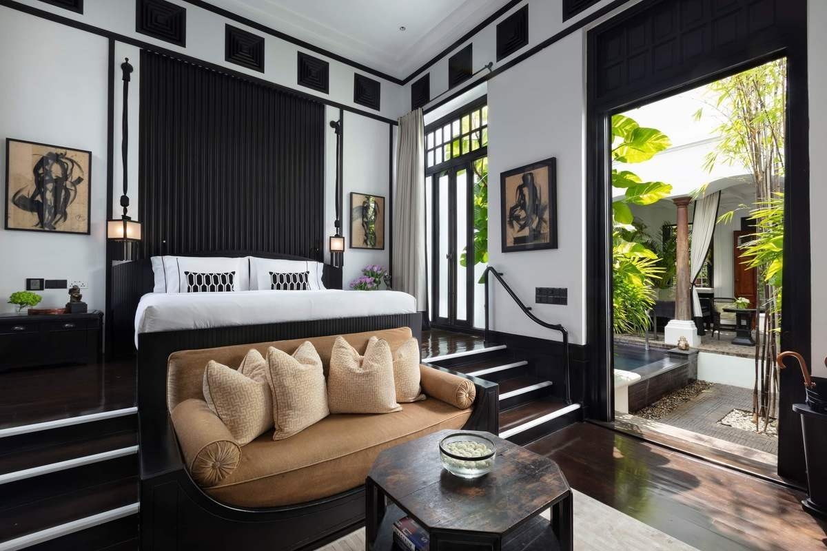 A high-ceilinged luxury bedroom at the Siam features a monochromatic Art Deco design with a raised bed area, tan sofa, and private courtyard access.