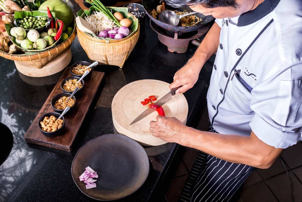 Overhead view of a chef in a white uniform slicing a red chili on a round wooden cutting board.