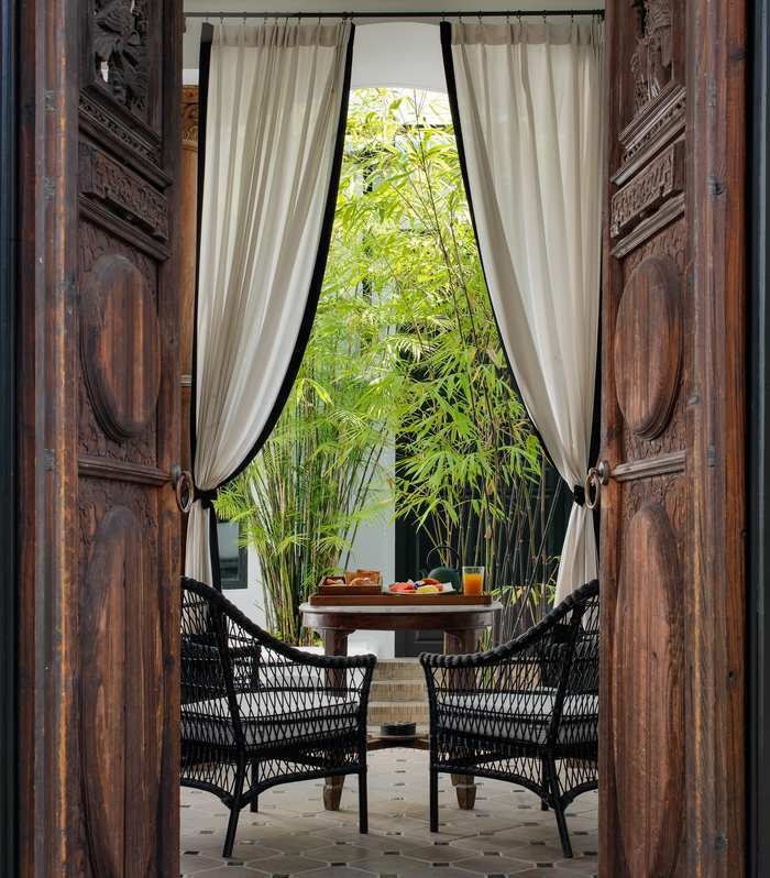 View through open ornate wooden doors of a small outdoor table with breakfast and wicker chairs.