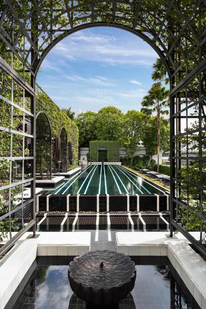 View through a garden archway of a long black and white striped swimming pool lined with green foliage.