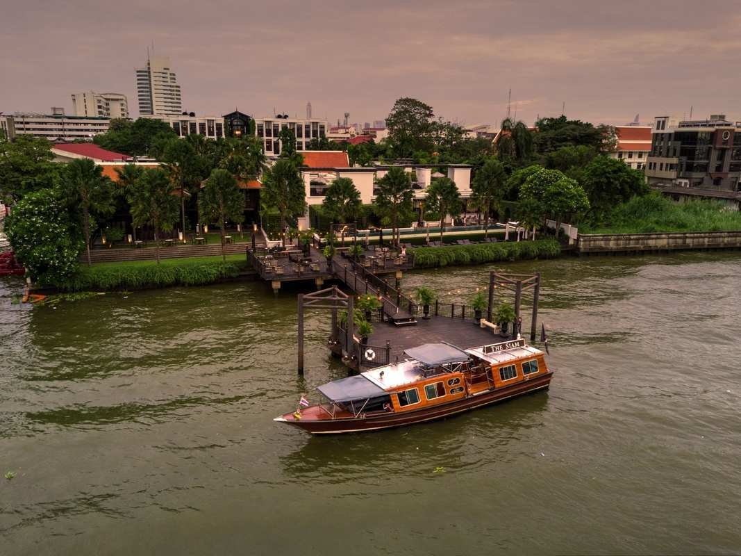 A wooden shuttle boat with The Siam text docked at a riverside pier with the city skyline in the background.