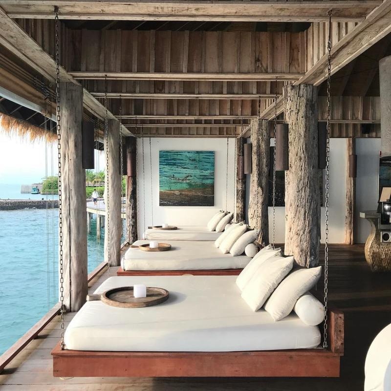 Three cushioned hanging beds on a shaded wooden deck facing the ocean with blue artwork on the wall.