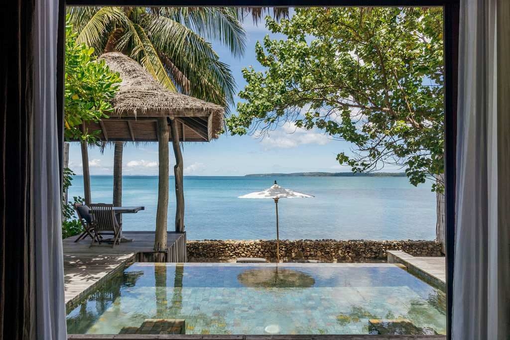 : View through a window, a thatched-roof hut and the blue sea under a bright sky.