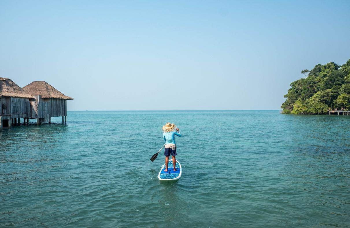 A person paddleboarding on clear blue water with tropical villas and a green island in the background.