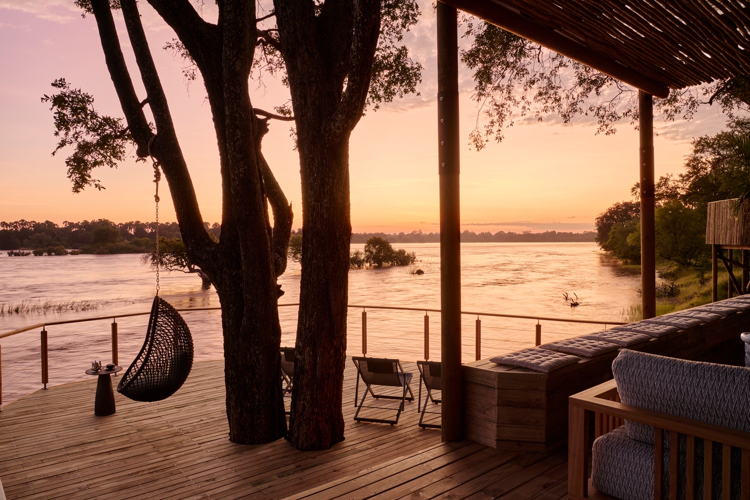 A hanging chair on a wooden deck during a pink sunset over the river at Victoria Falls River Lodge.