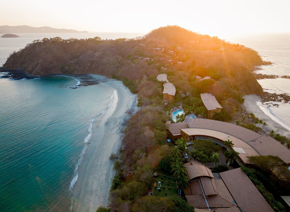 Aerial view of a tropical peninsula and resort buildings during a bright orange sunset.