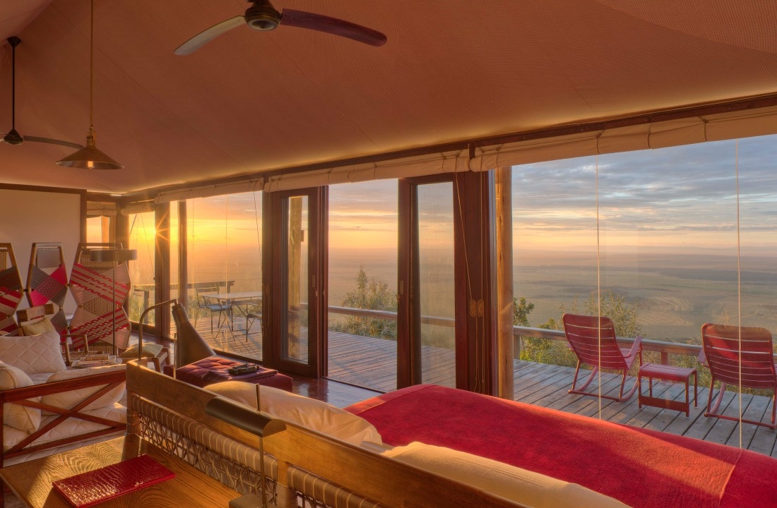 Luxury safari tent interior with a red bed and glass doors leading to a balcony with red rocking chairs at sunset.