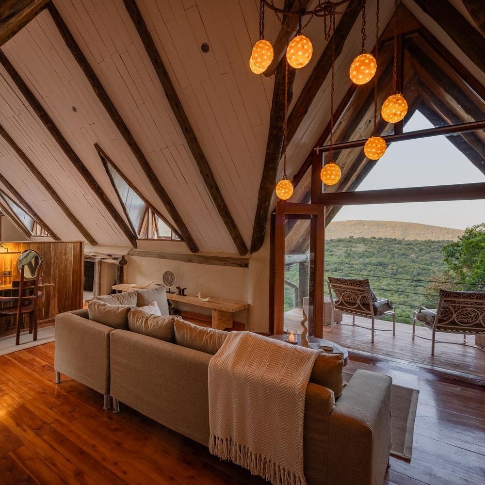 Spacious bedroom lounge with vaulted ceilings, exposed beams, a large sofa, and an open doorway to a deck overlooking a valley.