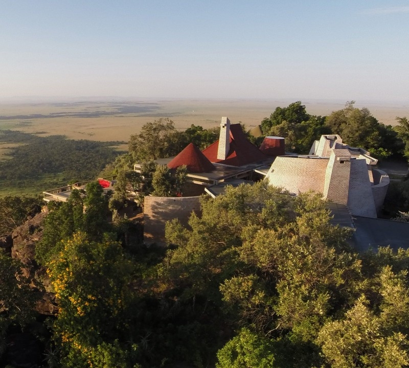 Aerial view of a hilltop lodge with conical red roofs surrounded by green trees overlooking a wide plain.