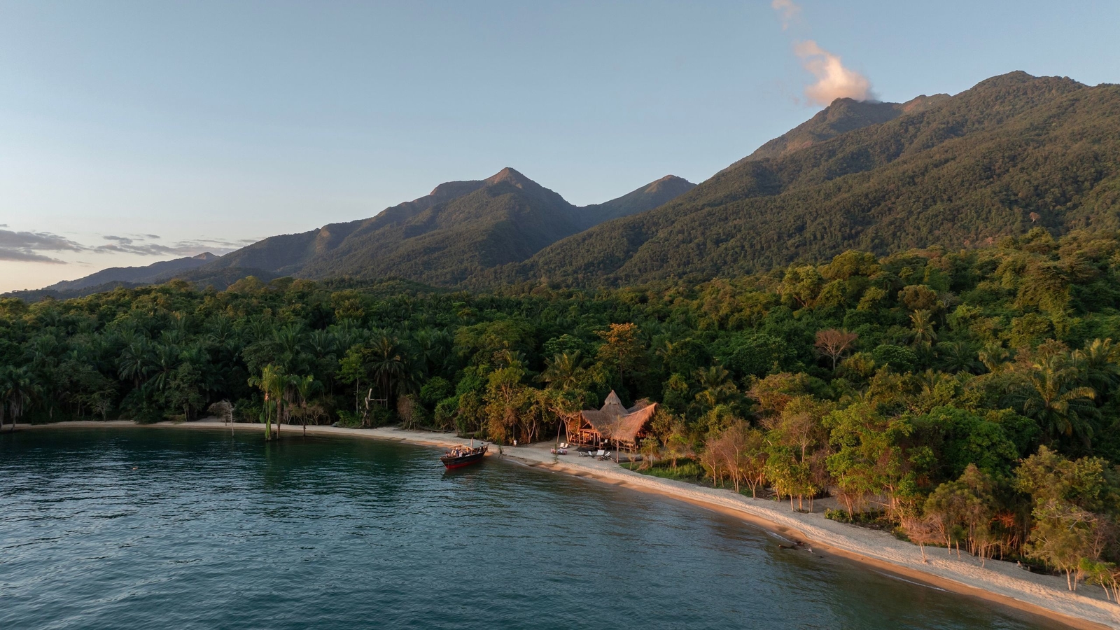 Aerial view of a remote beach lodge with thatched roofs on a white sand beach surrounded by dense jungle and mountains.