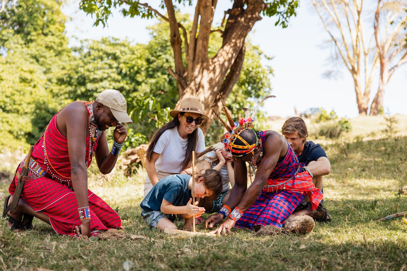 Maasai men demonstrating traditional fire-starting techniques with wood to children and adults outdoors.