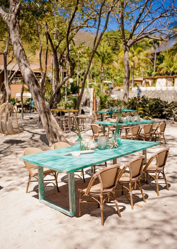 Outdoor dining area on a sandy beach with bright teal tables and tan chairs under leafy trees.