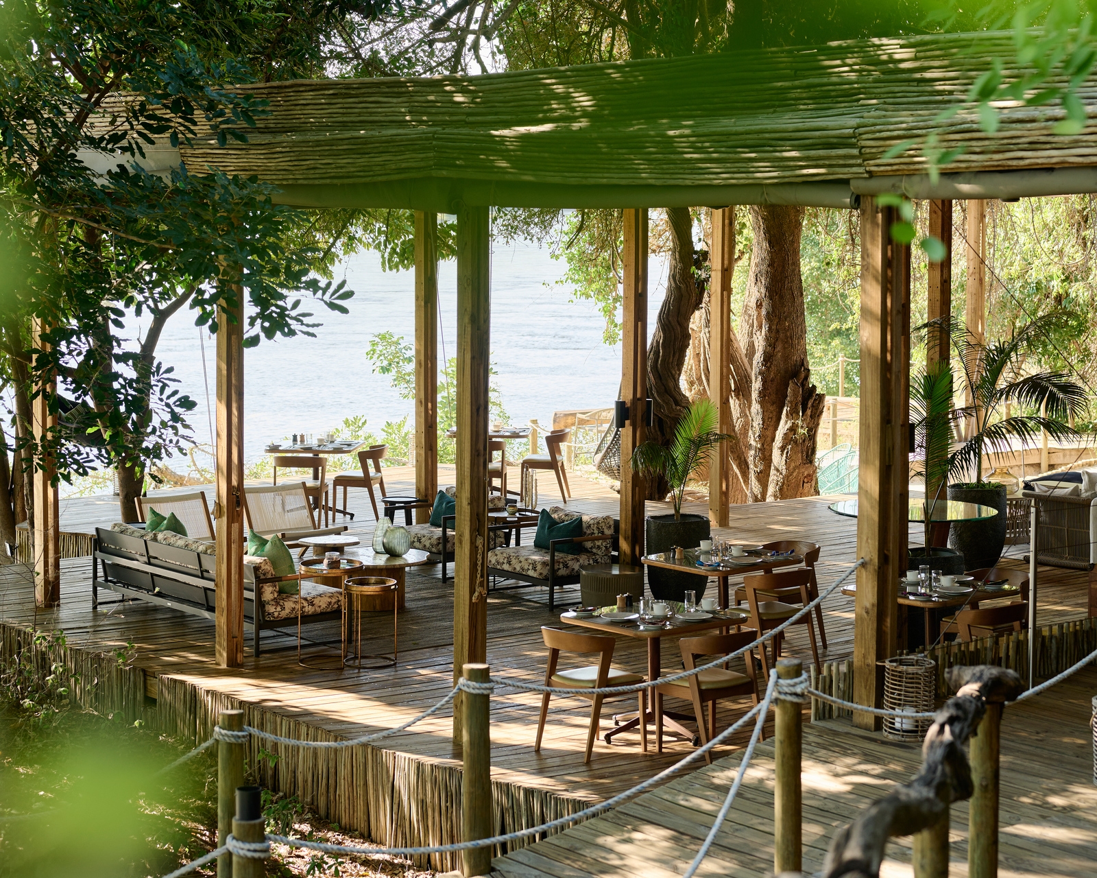 Outdoor dining area with wooden tables and chairs on a deck surrounded by trees at Victoria Falls River Lodge.