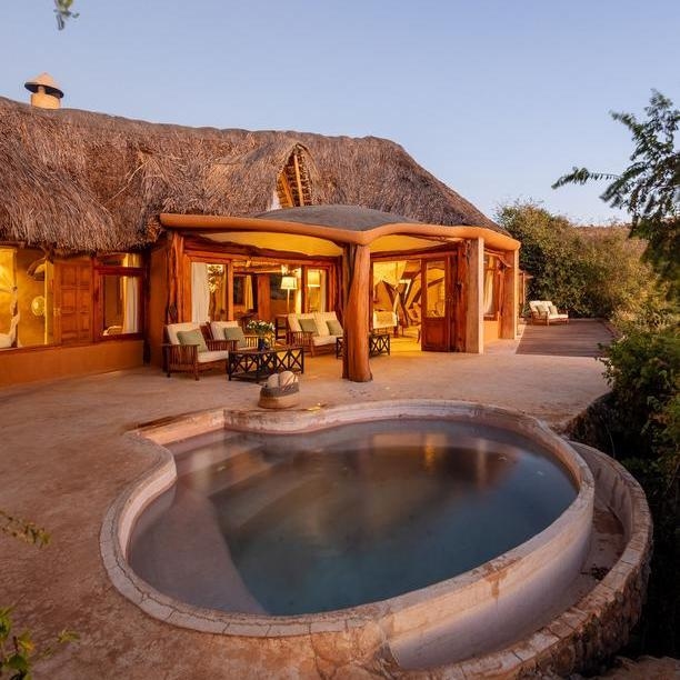 An outdoor plunge pool built into a stone patio in front of a thatched-roof lodge.