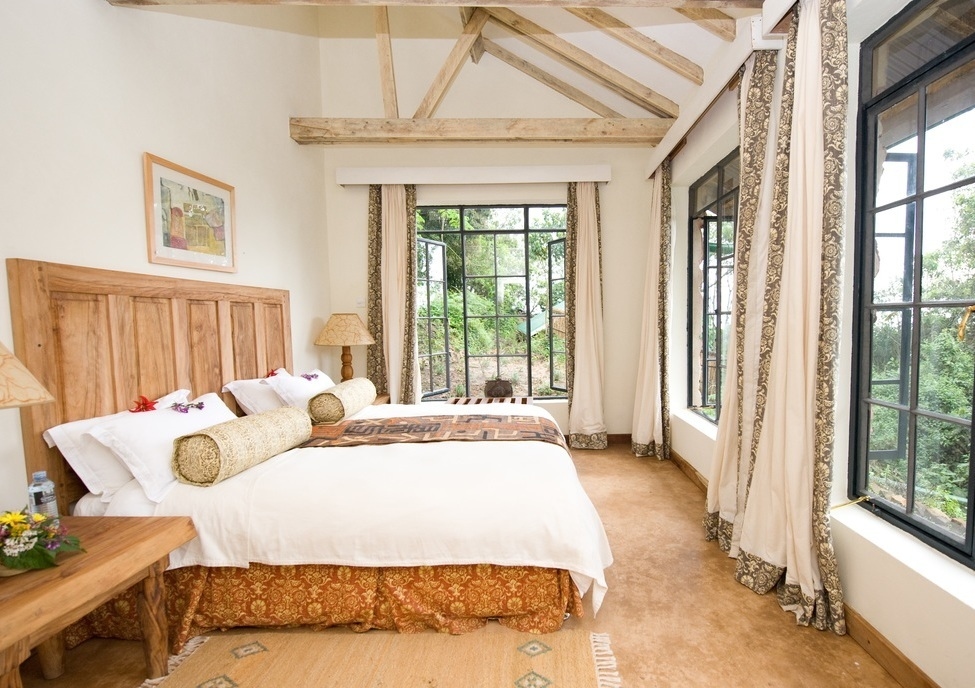 A bright bedroom with a wooden bed, exposed ceiling beams, and large black-framed windows overlooking green foliage.