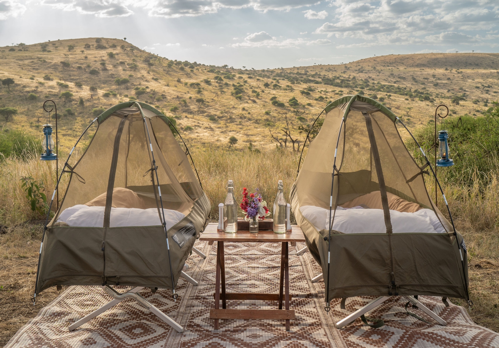 Two mesh tent cots arranged side by side outdoors with a small wooden table between them.