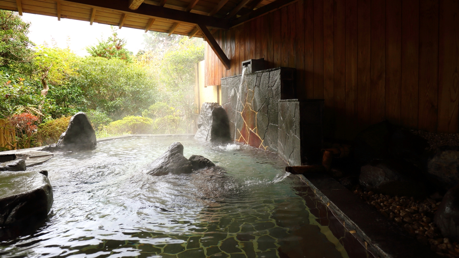 Steaming water in an outdoor stone hot spring pool under a wooden roof.