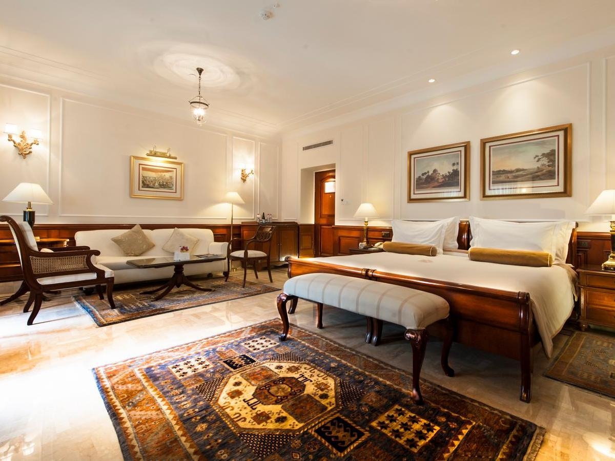 A luxury bedroom with a large bed, white sofa, patterned rugs, and framed art on white walls in a Grand Heritage Room.