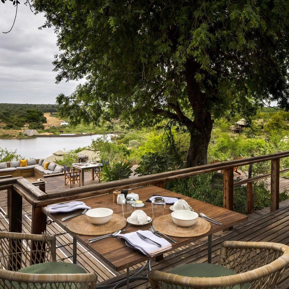 Breakfast table set for two on a wooden balcony overlooking a scenic river at Simbavati Hilltop Lodge.