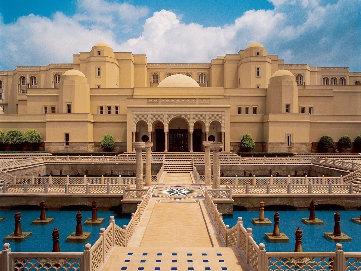 Symmetrical sandstone hotel exterior with tiered reflection pools and decorative fountains under a blue sky.