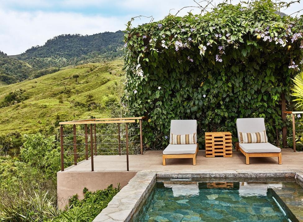 A stone plunge pool and two lounge chairs on a terrace overlooking a vast green mountain landscape.
