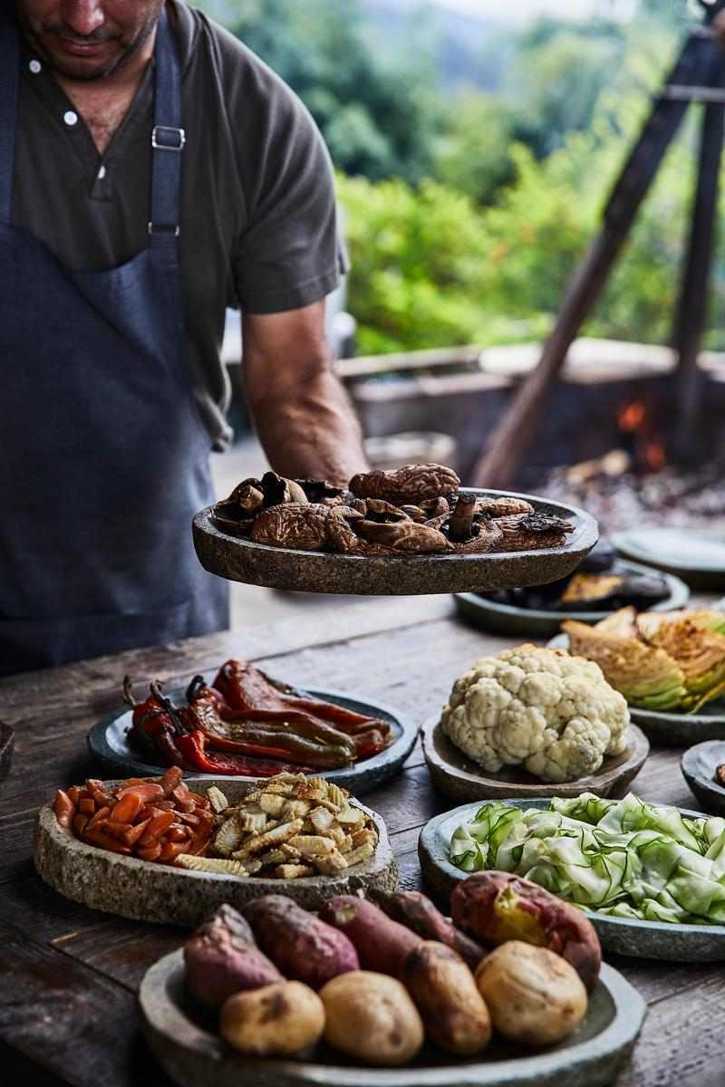 A chef holding a stone plate of roasted mushrooms next to dishes of cauliflower, peppers, and sweet potatoes.