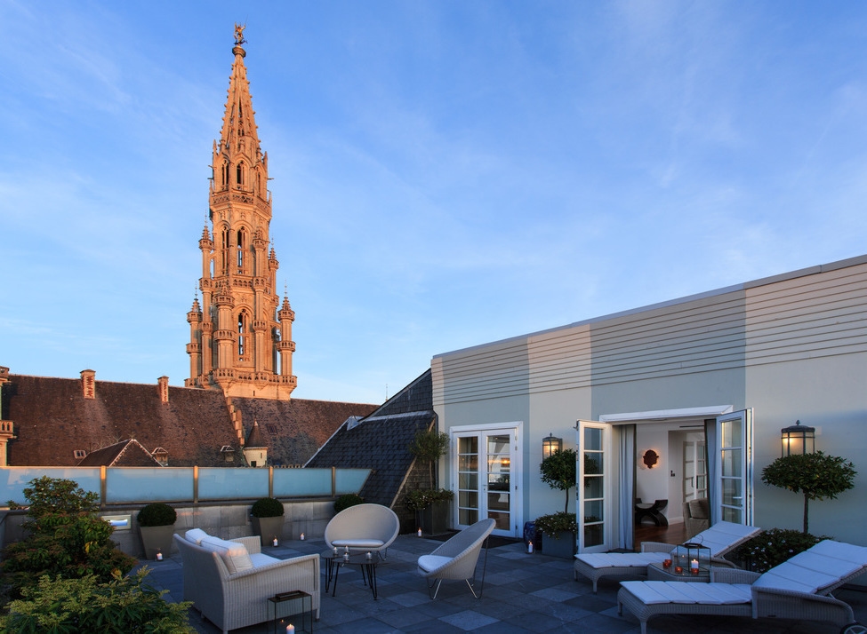 Rooftop terrace at Hotel Amigo Belgium with lounge seating, candles, and views of a tall gothic spire at dusk.