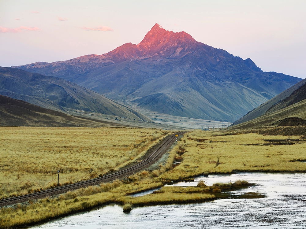 A glowing mountain peak at sunset overlooking railway tracks in a grassy valley with a stream.